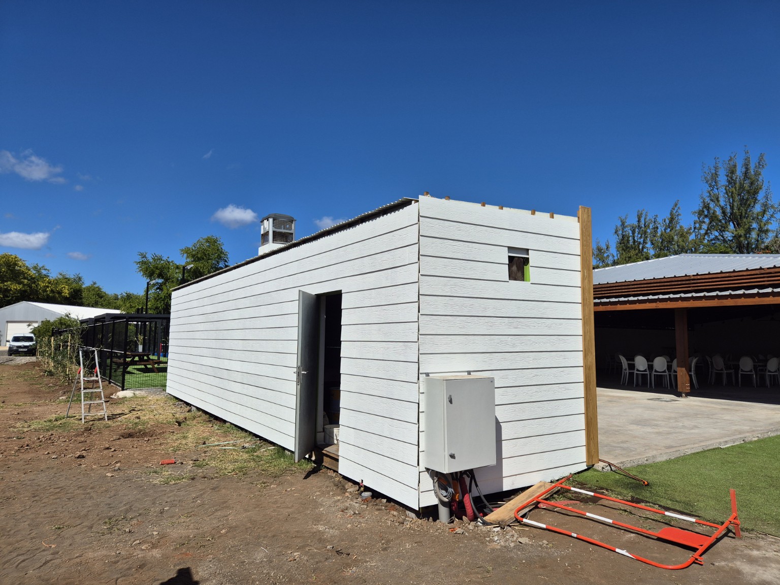 Deux façades du conteneur habillées de bardage fibres-ciment blanc, avec une porte ouverte et une petite ouverture en hauteur.
